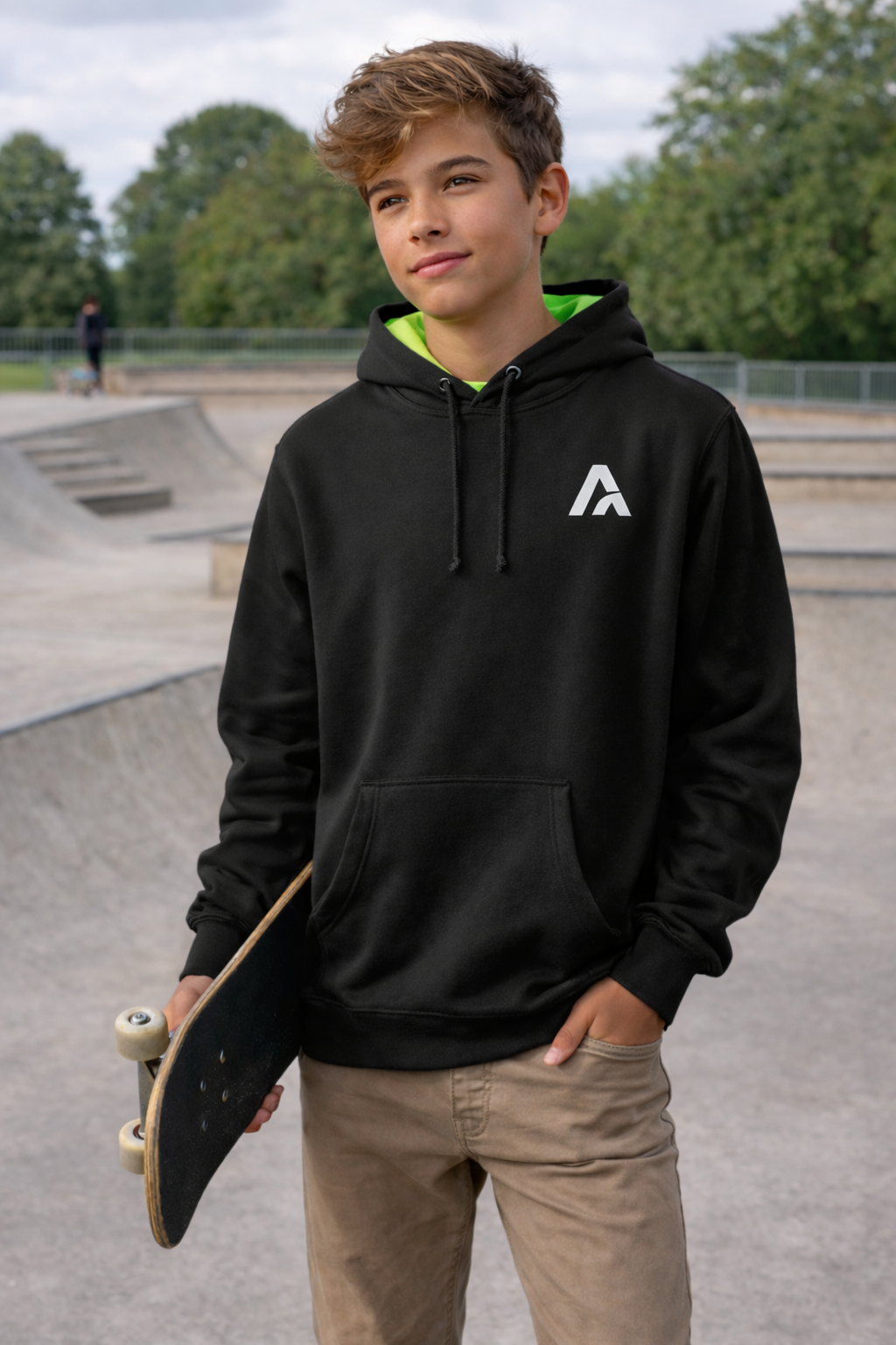 Young person wearing a black hoodie with a logo, holding a skateboard at a skate park.