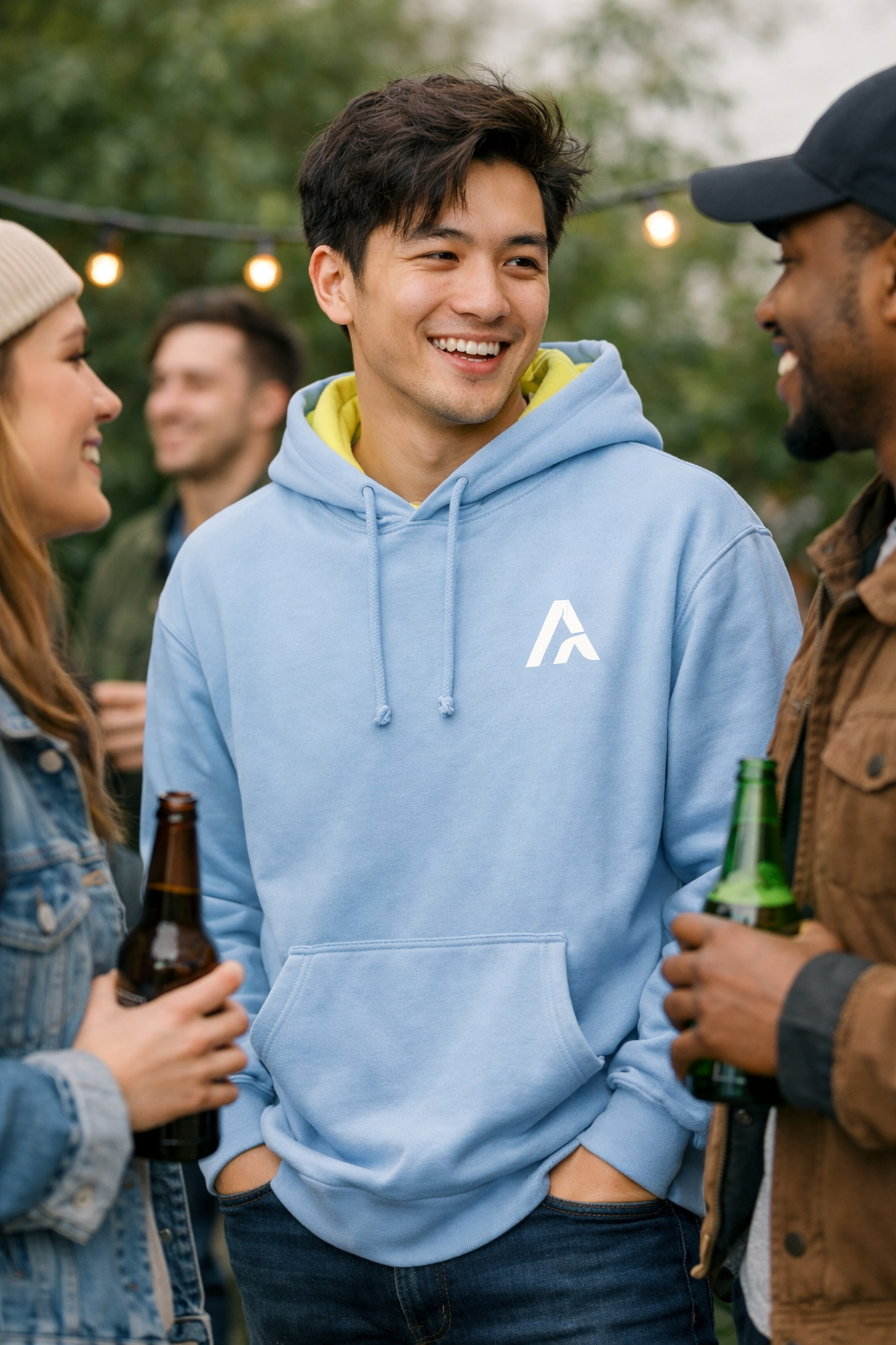 Man wearing a light blue hoodie with a logo, standing among friends at an outdoor gathering.
