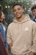 Man wearing a beige hoodie with a logo, standing outdoors with people in the background.