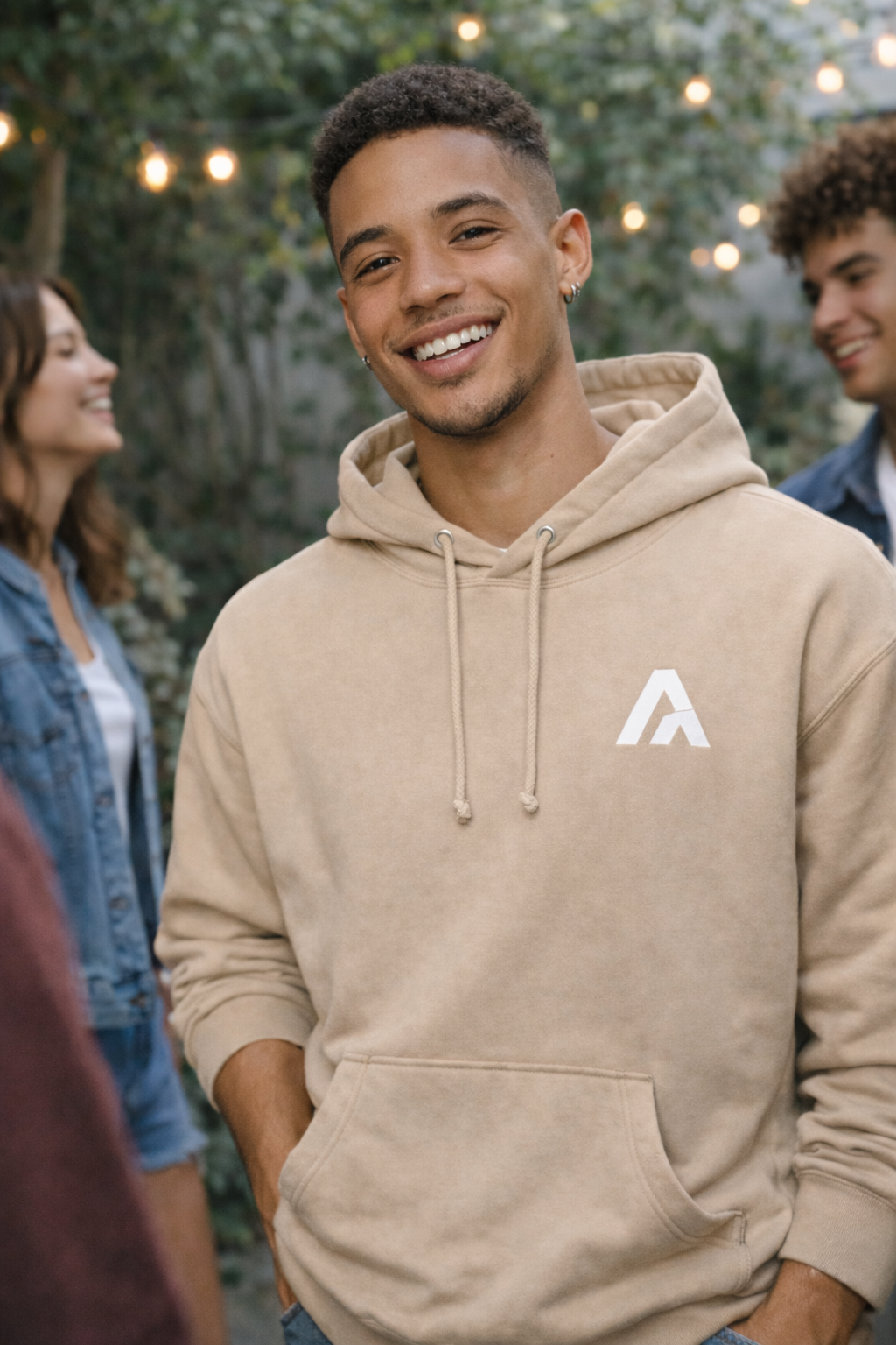 Man wearing a beige hoodie with a logo, standing outdoors with people in the background.