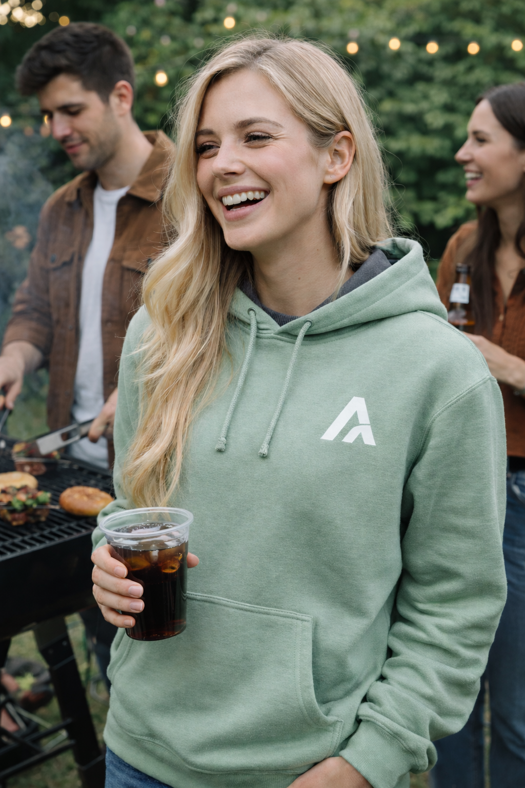 Woman in a green hoodie with a logo, holding a drink, standing outdoors with friends.