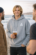 Man in a gray hoodie with a logo holding a drink on a beach