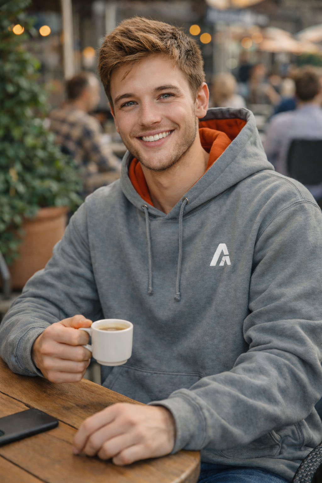 Man in a gray hoodie with a logo holding a coffee cup outdoors.
