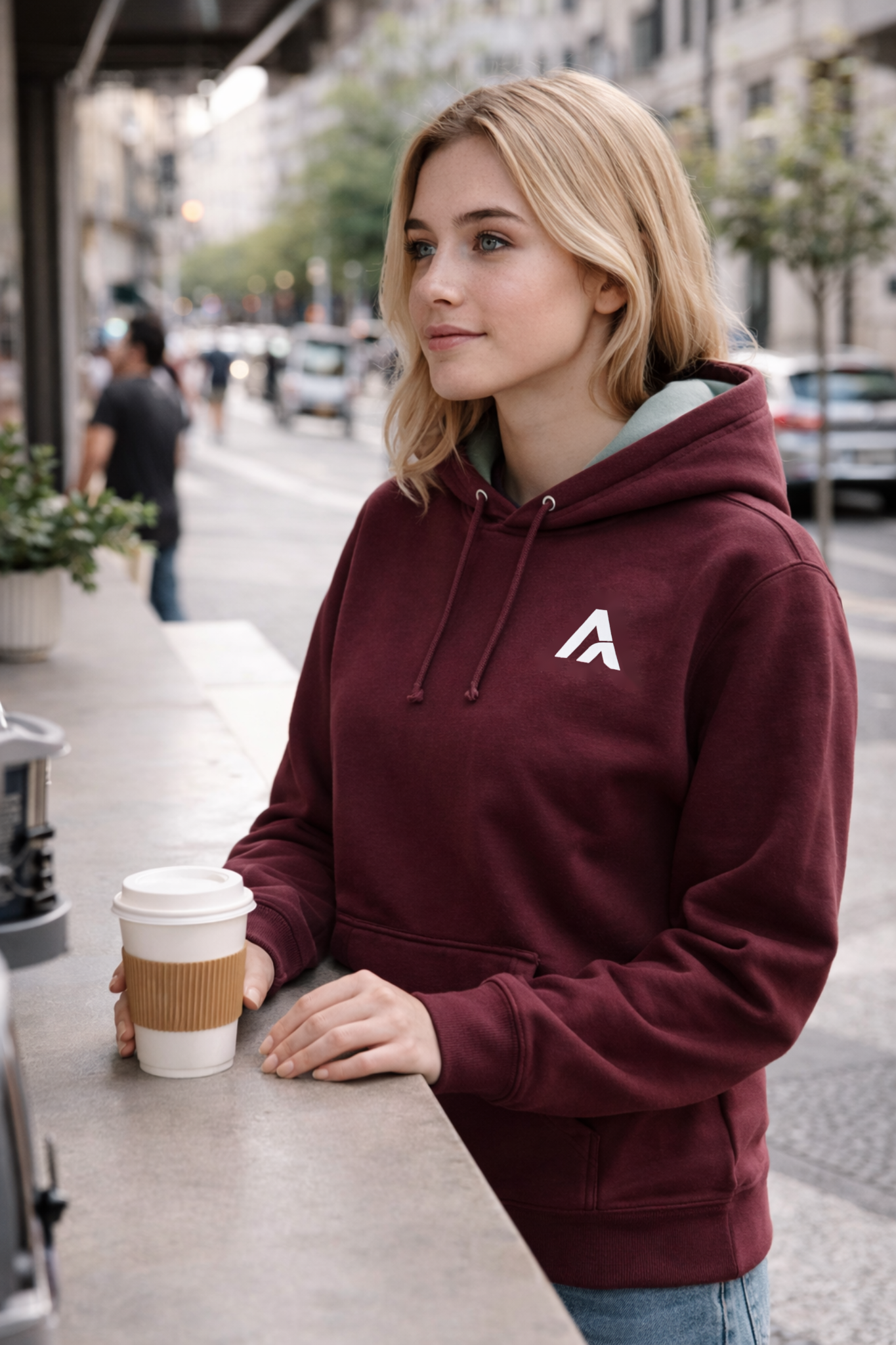 Woman in a maroon hoodie with a logo, holding a coffee cup on a city street.