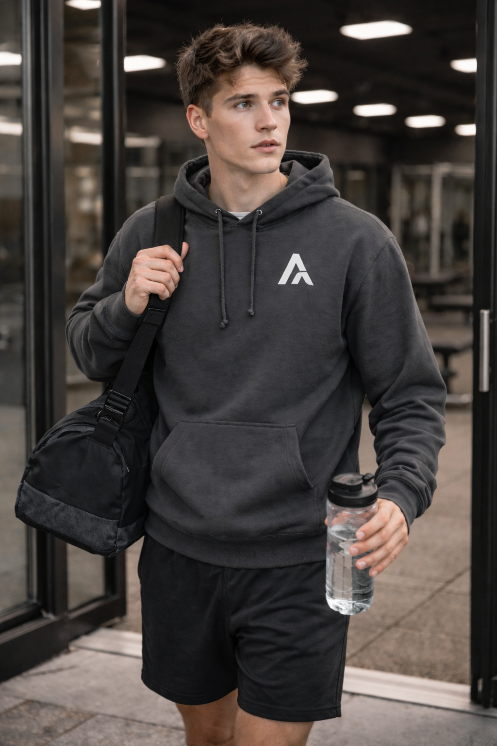 Man wearing a dark gray hoodie with a logo, holding a black duffel bag and water bottle, standing outside.