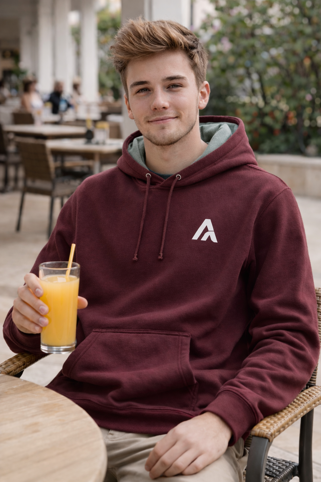 Person wearing a maroon hoodie with a logo, holding a glass of orange juice outdoors.