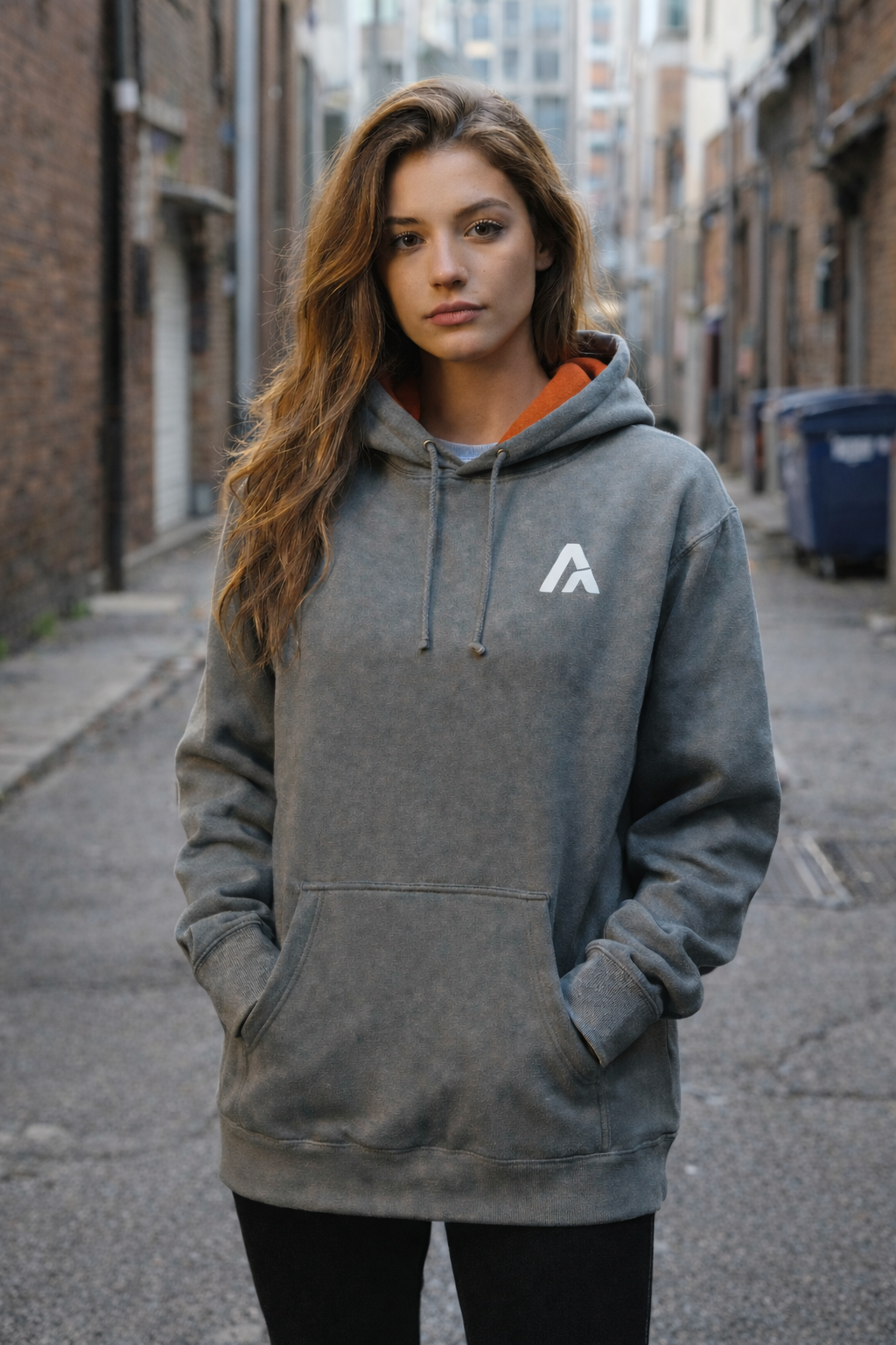 Female model wearing an Atlaswear heather grey premium hoodie with orange hood lining and minimalist embroidered logo, shown front-on in back alley setting