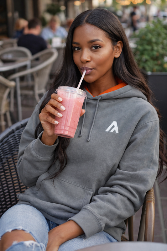 Female model wearing an Atlaswear heather grey premium hoodie with orange hood lining and minimalist embroidered logo, shown front-on drinking smoothie