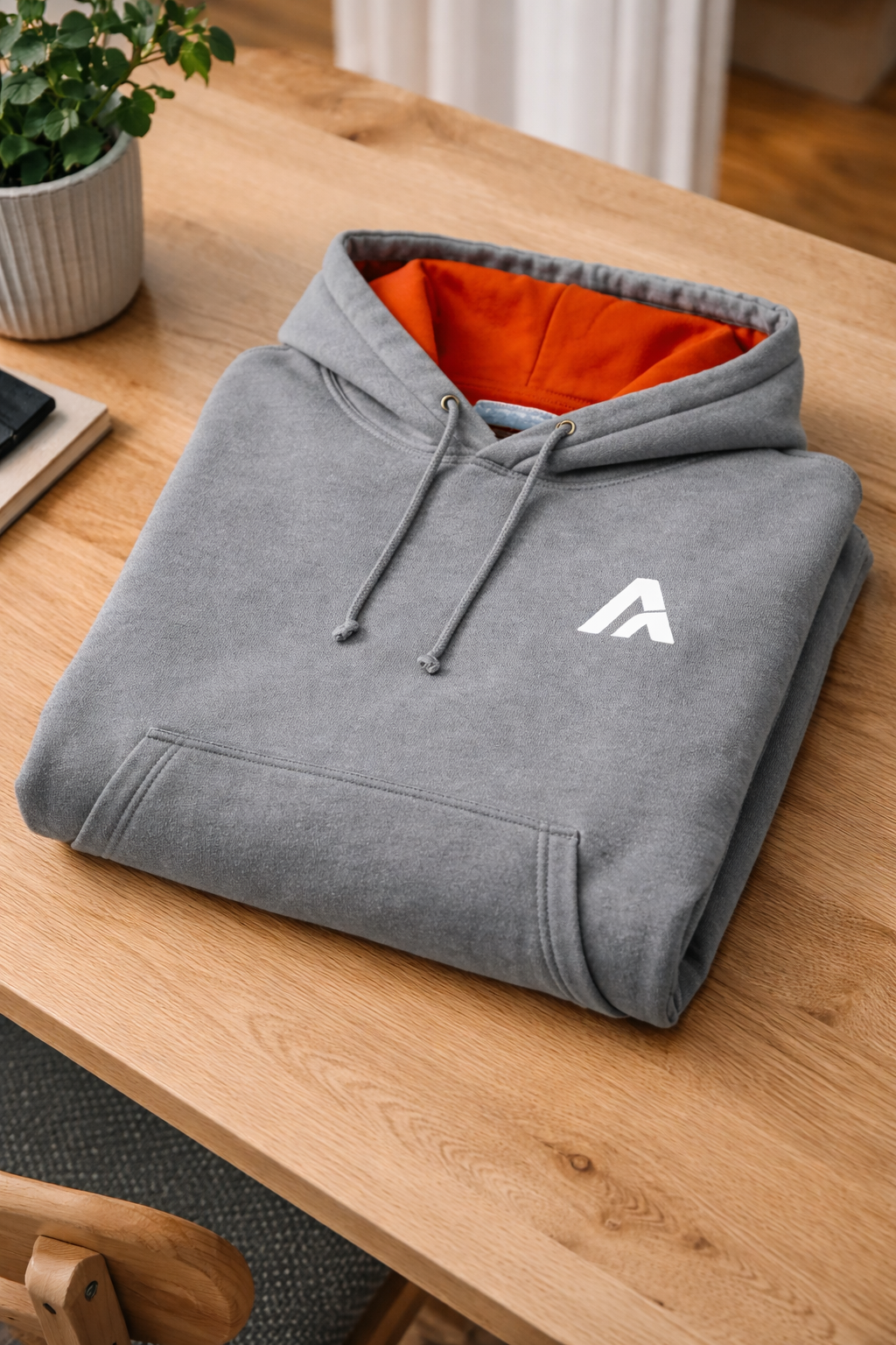 Atlaswear heather grey premium hoodie folded on a wooden table, showcasing the orange hood lining and minimalist embroidered logo in a clean lifestyle setting