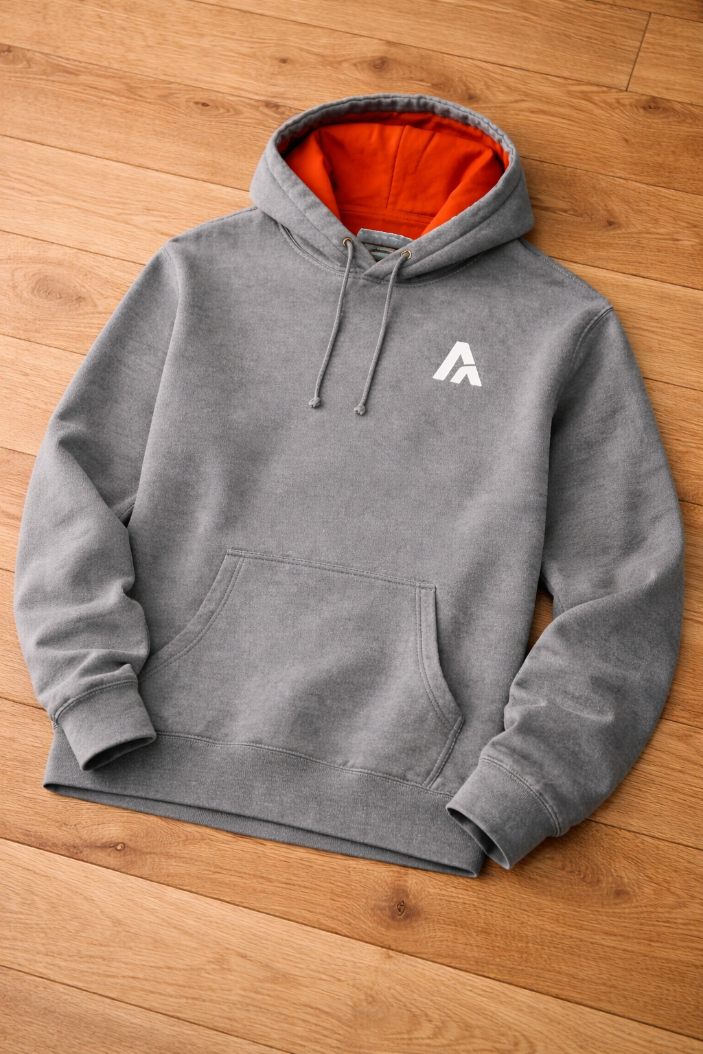 Atlaswear heather grey premium hoodie laid flat on a wooden floor, featuring a vibrant orange hood lining and minimalist embroidered logo for modern streetwear style