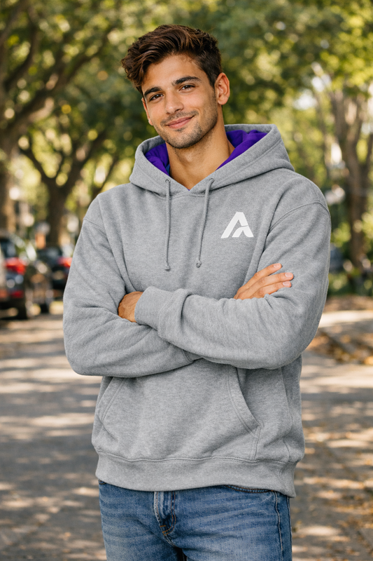 Male model wearing an Atlaswear heather grey premium hoodie with purple hood lining and embroidered logo, standing outdoors on a tree-lined street in a relaxed streetwear lifestyle look