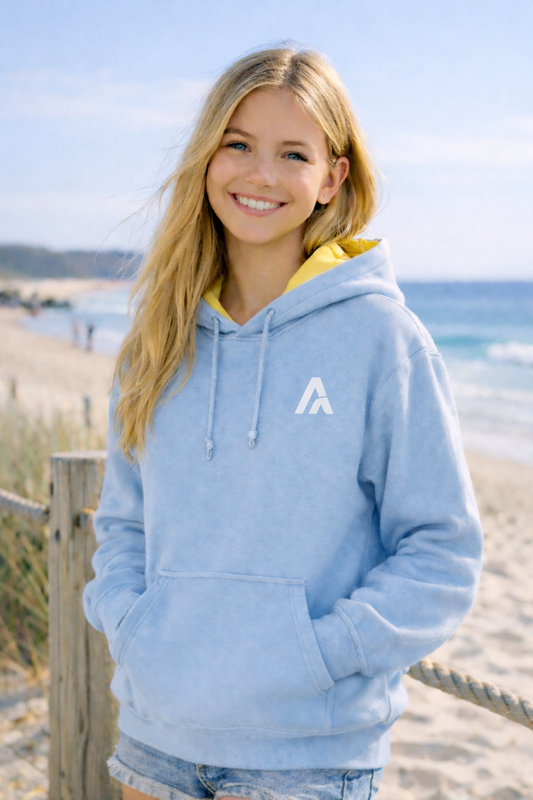 Female model wearing an Atlaswear light blue premium hoodie with yellow hood lining, standing by the beach in a relaxed coastal lifestyle setting