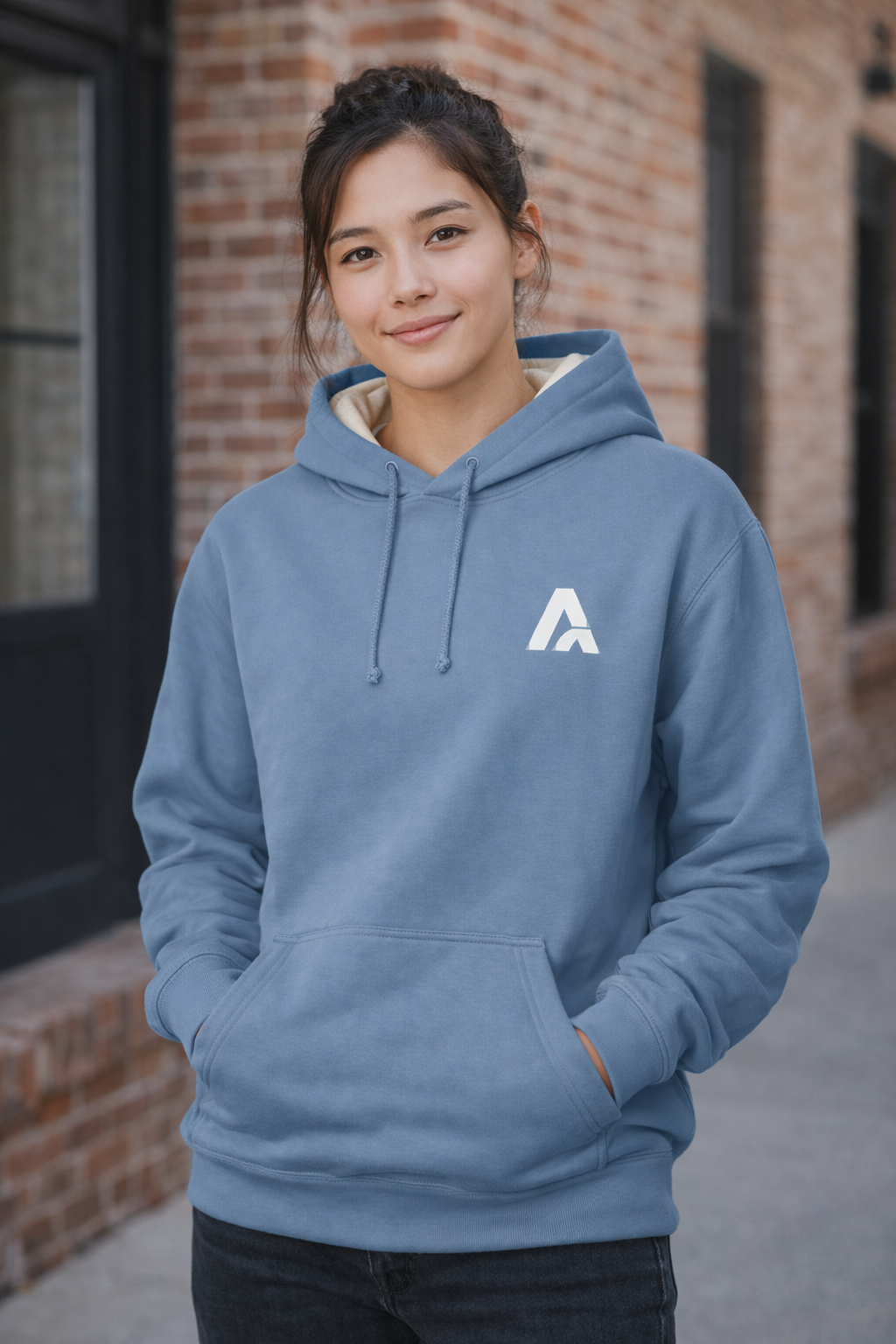Female model wearing an Atlaswear light blue premium hoodie with embroidered logo, standing outdoors against a brick wall in a relaxed streetwear lifestyle look