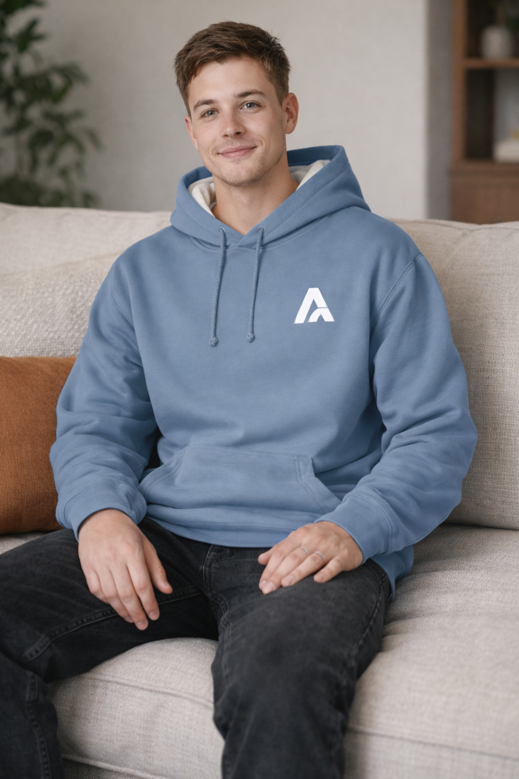 Male model wearing an Atlaswear light blue premium hoodie with embroidered logo, seated on a couch in a relaxed lifestyle setting showcasing everyday streetwear comfort